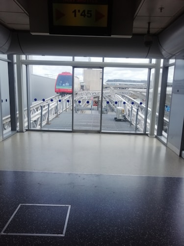 A monorail with a bright red front is visible through large glass windows on an elevated track. The indoor area features a tiled floor and some overhead signage indicating the shuttle's timing. The railing and the glass have a clean, modern design, and outside, there are cloudy skies visible.