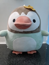 A plush toy resembling a penguin with a brown head, white face, a large pink beak, and a round white belly. It has small black eyes and is wearing a small yellow bow on its head. The plush appears soft and cuddly, sitting on a dark granite surface with a light blue and white background.