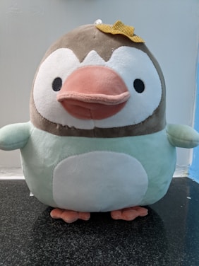 A plush toy resembling a penguin with a brown head, white face, a large pink beak, and a round white belly. It has small black eyes and is wearing a small yellow bow on its head. The plush appears soft and cuddly, sitting on a dark granite surface with a light blue and white background.