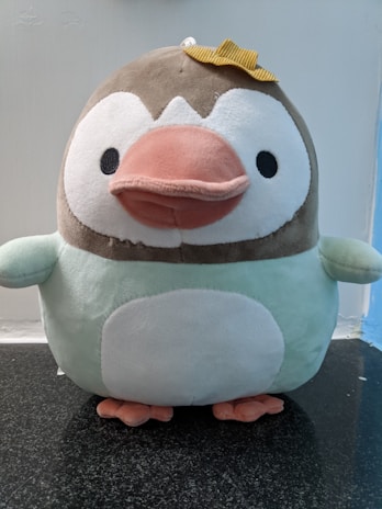A plush toy resembling a penguin with a brown head, white face, a large pink beak, and a round white belly. It has small black eyes and is wearing a small yellow bow on its head. The plush appears soft and cuddly, sitting on a dark granite surface with a light blue and white background.