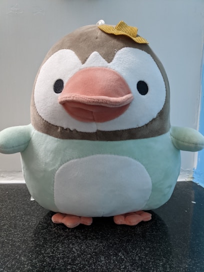 A plush toy resembling a penguin with a brown head, white face, a large pink beak, and a round white belly. It has small black eyes and is wearing a small yellow bow on its head. The plush appears soft and cuddly, sitting on a dark granite surface with a light blue and white background.