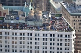 A rooftop gathering on a building in an urban setting features multiple tables with umbrellas, numerous people seated and interacting, and large industrial units on the adjacent rooftop area. The building showcases classic architectural elements, and the surroundings include a mix of historic and modern structures.