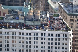 A rooftop gathering on a building in an urban setting features multiple tables with umbrellas, numerous people seated and interacting, and large industrial units on the adjacent rooftop area. The building showcases classic architectural elements, and the surroundings include a mix of historic and modern structures.