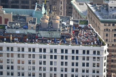 A rooftop gathering on a building in an urban setting features multiple tables with umbrellas, numerous people seated and interacting, and large industrial units on the adjacent rooftop area. The building showcases classic architectural elements, and the surroundings include a mix of historic and modern structures.