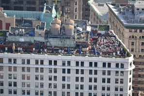 A rooftop gathering on a building in an urban setting features multiple tables with umbrellas, numerous people seated and interacting, and large industrial units on the adjacent rooftop area. The building showcases classic architectural elements, and the surroundings include a mix of historic and modern structures.