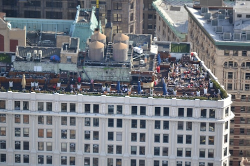 A rooftop gathering on a building in an urban setting features multiple tables with umbrellas, numerous people seated and interacting, and large industrial units on the adjacent rooftop area. The building showcases classic architectural elements, and the surroundings include a mix of historic and modern structures.