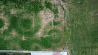 An aerial view of a sports field with goal posts, surrounded by patches of green grass and some areas that appear worn or bare. A sidewalk or concrete path runs along the bottom edge of the image, partially visible, with fencing sections visible throughout.