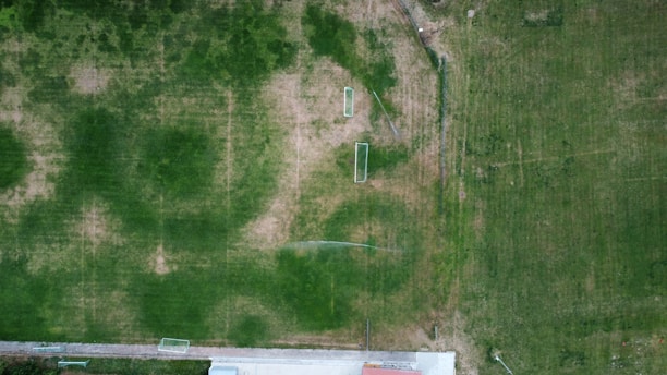 An aerial view of a sports field with goal posts, surrounded by patches of green grass and some areas that appear worn or bare. A sidewalk or concrete path runs along the bottom edge of the image, partially visible, with fencing sections visible throughout.