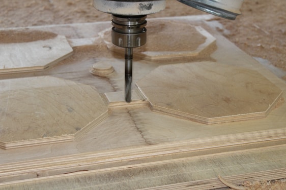 A CNC machine is actively carving out shapes from a sheet of plywood. The machining process involves a rotating cutting tool, creating precise geometric designs. Wood shavings are scattered around the area, indicating an ongoing task.