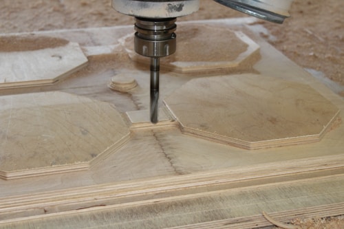 A CNC machine is actively carving out shapes from a sheet of plywood. The machining process involves a rotating cutting tool, creating precise geometric designs. Wood shavings are scattered around the area, indicating an ongoing task.