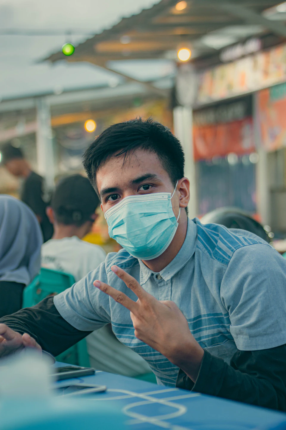 In the image, a young man is seated at a table outdoors, wearing a face mask and making a peace sign with his fingers. The setting appears to be an outdoor market or food court, with other people present but not visible in the frame.