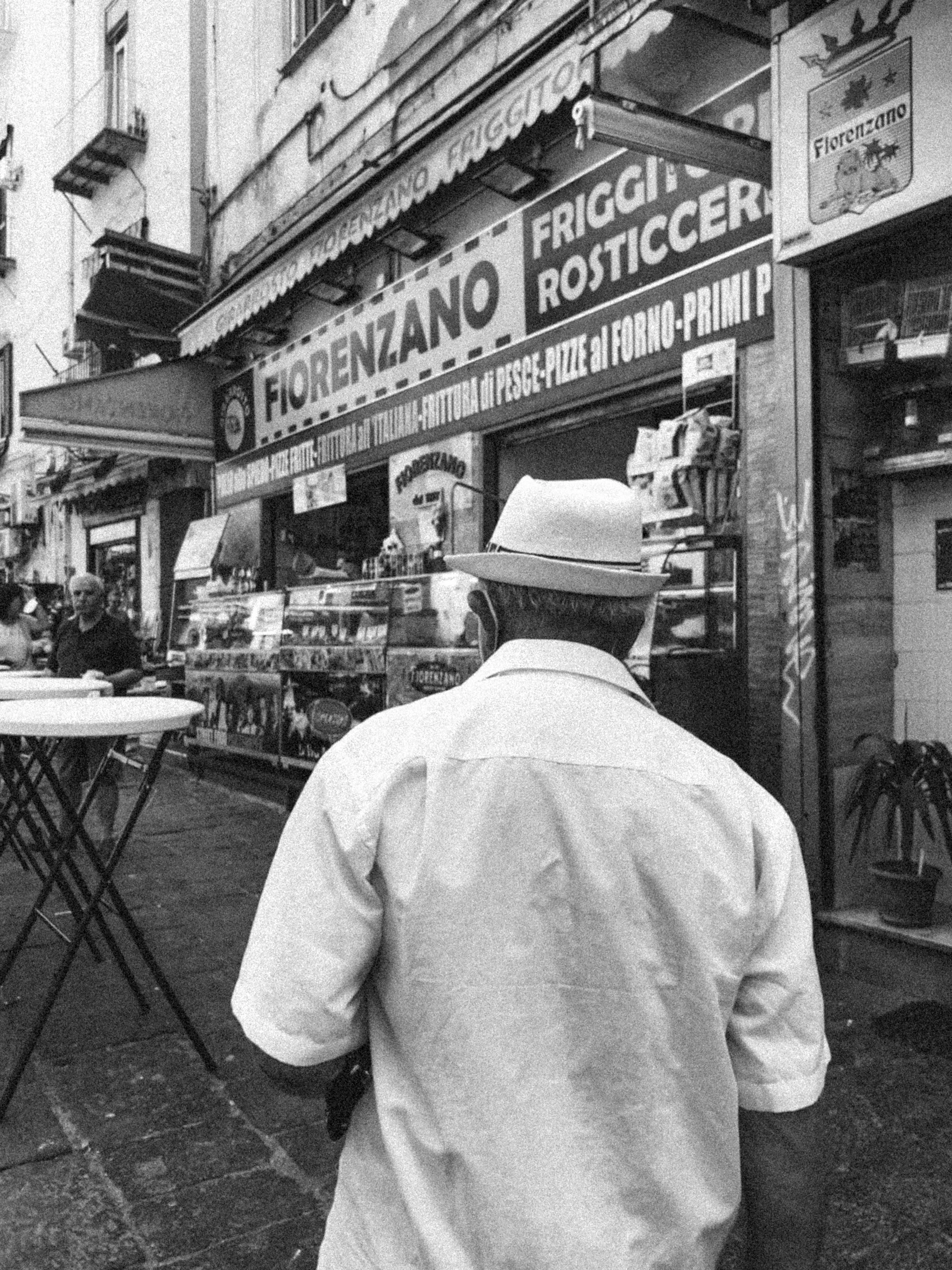 Black-and-white street scene of a man in a hat walking past Naples' Fiorenzano food shop.