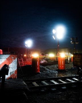 A nighttime construction site is illuminated by bright floodlights. The area includes traffic cones, metal fencing, and construction materials. A railway track is visible in the foreground, with a partially built structure nearby. There are warning signs indicating restricted access.