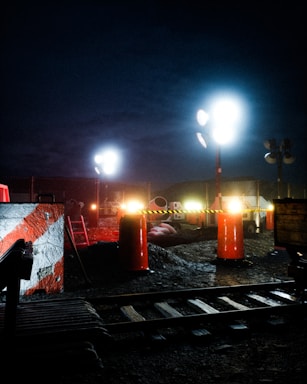 A nighttime construction site is illuminated by bright floodlights. The area includes traffic cones, metal fencing, and construction materials. A railway track is visible in the foreground, with a partially built structure nearby. There are warning signs indicating restricted access.
