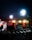 A nighttime construction site is illuminated by bright floodlights. The area includes traffic cones, metal fencing, and construction materials. A railway track is visible in the foreground, with a partially built structure nearby. There are warning signs indicating restricted access.