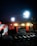 A nighttime construction site is illuminated by bright floodlights. The area includes traffic cones, metal fencing, and construction materials. A railway track is visible in the foreground, with a partially built structure nearby. There are warning signs indicating restricted access.