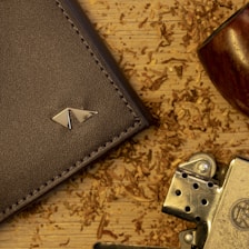 A close-up view featuring a brown leather wallet with a metallic logo, a metal lighter, and a wooden pipe on a wooden surface scattered with tobacco shavings.