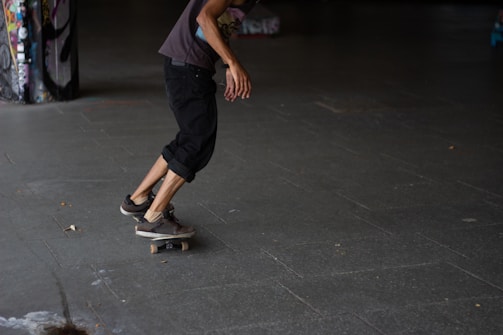 A person is skateboarding on a smooth, dark surface. They are dressed casually in a t-shirt and rolled-up pants. The space appears to be an urban setting with graffiti on a nearby pillar, suggesting a skate park or street environment.