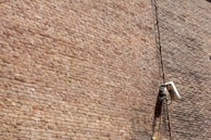 A textured brick wall with a mounted surveillance camera and a cable running vertically alongside it.