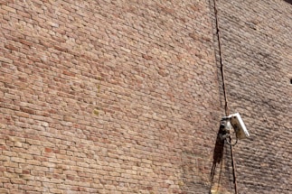 A textured brick wall with a mounted surveillance camera and a cable running vertically alongside it.
