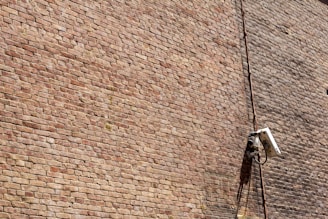 A textured brick wall with a mounted surveillance camera and a cable running vertically alongside it.