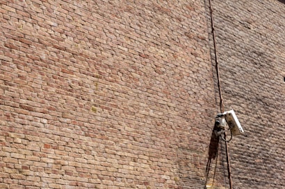 A textured brick wall with a mounted surveillance camera and a cable running vertically alongside it.