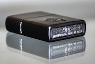 A close-up view of a metallic lighter with engraved details, showing the brand name and place of manufacture. It rests on a reflective surface, creating a mirror image effect underneath.