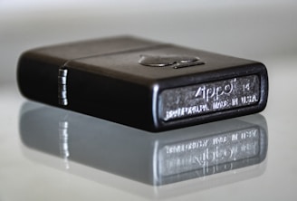 A close-up view of a metallic lighter with engraved details, showing the brand name and place of manufacture. It rests on a reflective surface, creating a mirror image effect underneath.