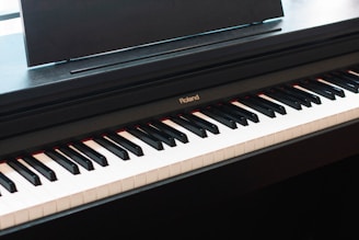 A digital piano with black and white keys is shown, prominently displaying the 'Roland' brand above the keyboard. The piano has a sleek design with a black finish, and a music stand attached.