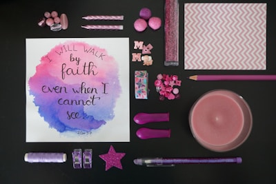 A variety of pink and purple stationery and decorative items arranged around a piece of artwork with a motivational quote. The artwork has watercolor accents in pink and purple, and the quote reads 'I will walk by faith even when I cannot see'. The items include buttons, candles, pom-poms, a star decoration, glitter, and a pencil.