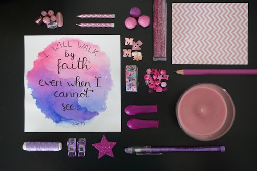 A variety of pink and purple stationery and decorative items arranged around a piece of artwork with a motivational quote. The artwork has watercolor accents in pink and purple, and the quote reads 'I will walk by faith even when I cannot see'. The items include buttons, candles, pom-poms, a star decoration, glitter, and a pencil.