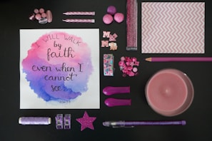 A variety of pink and purple stationery and decorative items arranged around a piece of artwork with a motivational quote. The artwork has watercolor accents in pink and purple, and the quote reads 'I will walk by faith even when I cannot see'. The items include buttons, candles, pom-poms, a star decoration, glitter, and a pencil.