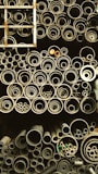 A collection of pipes stored in an organized manner, ranging from large to small diameters. The pipes are stacked vertically and closely packed, giving a sense of industrial storage. A metal ladder is positioned on the left side, partially visible.