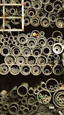 A collection of pipes stored in an organized manner, ranging from large to small diameters. The pipes are stacked vertically and closely packed, giving a sense of industrial storage. A metal ladder is positioned on the left side, partially visible.