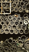 A collection of pipes stored in an organized manner, ranging from large to small diameters. The pipes are stacked vertically and closely packed, giving a sense of industrial storage. A metal ladder is positioned on the left side, partially visible.