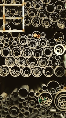 A collection of pipes stored in an organized manner, ranging from large to small diameters. The pipes are stacked vertically and closely packed, giving a sense of industrial storage. A metal ladder is positioned on the left side, partially visible.