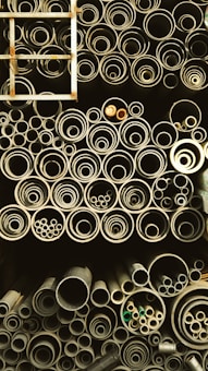 A collection of pipes stored in an organized manner, ranging from large to small diameters. The pipes are stacked vertically and closely packed, giving a sense of industrial storage. A metal ladder is positioned on the left side, partially visible.