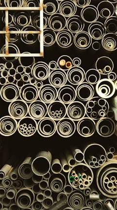 A collection of pipes stored in an organized manner, ranging from large to small diameters. The pipes are stacked vertically and closely packed, giving a sense of industrial storage. A metal ladder is positioned on the left side, partially visible.