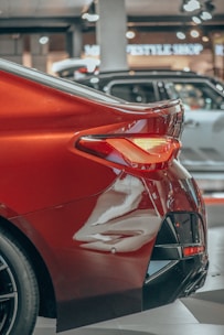 Close-up view of the rear section of a shiny red car with visible taillight and part of the bumper. In the background, there are a few other cars in a showroom setting with the storefront signs slightly out of focus.