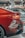 Close-up view of the rear section of a shiny red car with visible taillight and part of the bumper. In the background, there are a few other cars in a showroom setting with the storefront signs slightly out of focus.