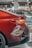 Close-up view of the rear section of a shiny red car with visible taillight and part of the bumper. In the background, there are a few other cars in a showroom setting with the storefront signs slightly out of focus.