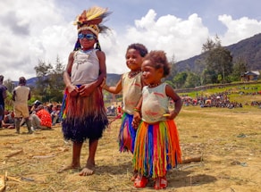 Three children stand outdoors wearing traditional attire and colorful skirts. The scene is lively with people gathered in the background, and the landscape includes hills and scattered trees under a partly cloudy sky.