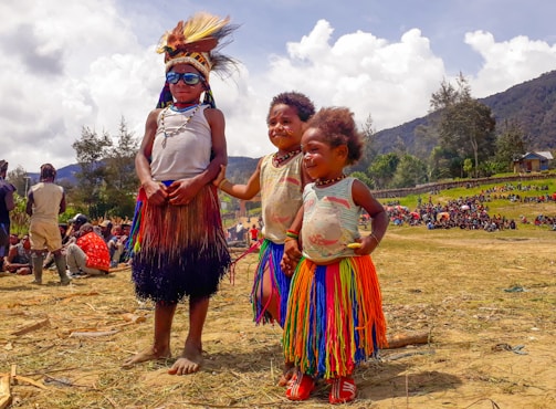 Three children stand outdoors wearing traditional attire and colorful skirts. The scene is lively with people gathered in the background, and the landscape includes hills and scattered trees under a partly cloudy sky.