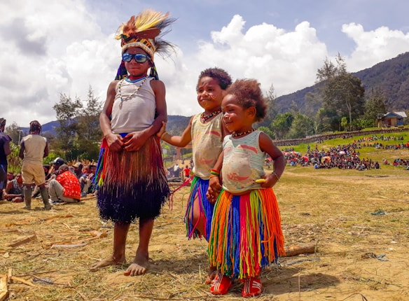 Three children stand outdoors wearing traditional attire and colorful skirts. The scene is lively with people gathered in the background, and the landscape includes hills and scattered trees under a partly cloudy sky.