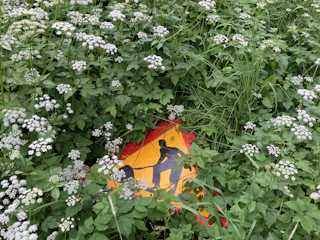 A road sign partially obscured by dense green foliage and white wildflowers, depicting a construction worker. The sign is predominantly red and yellow, with black silhouettes.