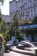 A city park features a decorative fountain with water spouting into a pool. There is lush greenery and flowers surrounding the area. A carousel with a colorful, ornate roof is visible in the background, and a large building with numerous windows and balconies looms nearby. A bench is placed in the foreground with a person seated, partially obscured by foliage.