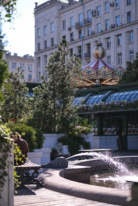 A city park features a decorative fountain with water spouting into a pool. There is lush greenery and flowers surrounding the area. A carousel with a colorful, ornate roof is visible in the background, and a large building with numerous windows and balconies looms nearby. A bench is placed in the foreground with a person seated, partially obscured by foliage.