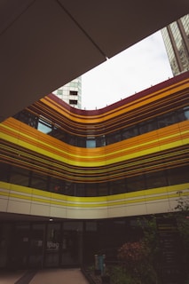 A modern building with a facade featuring a series of horizontal stripes in shades of yellow and orange. The structure stands in a courtyard with high-rise buildings visible in the background. The bottom part of the building has large glass windows and there are plants and signage visible near the entrance.
