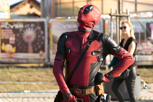 A person in a red and black superhero costume poses confidently with hands on hips in an outdoor setting. The background features blurred banners and a person wearing sunglasses standing casually.