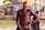 A person in a red and black superhero costume poses confidently with hands on hips in an outdoor setting. The background features blurred banners and a person wearing sunglasses standing casually.
